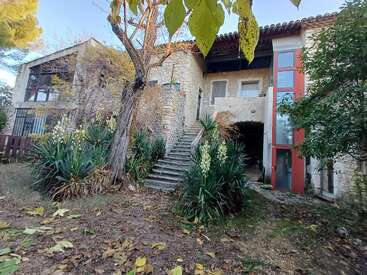 Cette image montre une maison rustique en pierre avec des escaliers extérieurs, entourée d'une végétation luxuriante, de grandes plantes à feuilles et d'arbres. Le cadre semble calme et naturel.