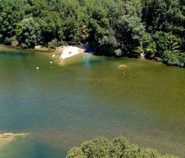 Une scène au bord d'une rivière avec une petite plage de sable, des gens qui se baignent, entourés d'arbres verdoyants, et un village à flanc de colline avec un carré bleu soulignant les maisons.