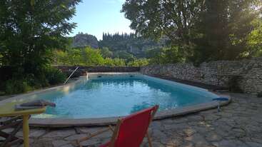 Ein ruhiger Pool mit klarem, blauem Wasser, umgeben von Steinpflaster, üppigem Grün und einer Steinmauer, mit Hügeln und Zypressen im Hintergrund.
