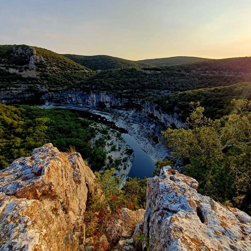 Des falaises rocheuses dentelées surplombent une rivière sinueuse en contrebas, entourée d'une forêt verte dense et de collines ondulantes. La lumière douce du soleil baigne le paysage paisible au coucher du soleil.
