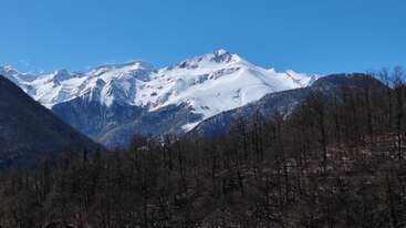 Un impresionante paisaje montañoso muestra picos nevados bajo un cielo azul y despejado, con árboles sin hojas y colinas escarpadas en primer plano que aportan textura y profundidad.