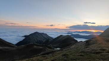 La imagen muestra un impresionante paisaje montañoso al amanecer, con colinas onduladas, nubes que cubren los valles y la cálida luz del sol asomándose por el horizonte lejano. Tranquilo y pintoresco.