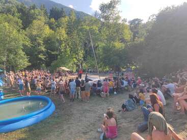 Un numeroso grupo de personas se reúne al aire libre en una zona boscosa para asistir a una actuación en el centro del escenario. Unas piscinas hinchables azules y frondosos árboles verdes rodean al público, que disfruta del momento con total tranquilidad.