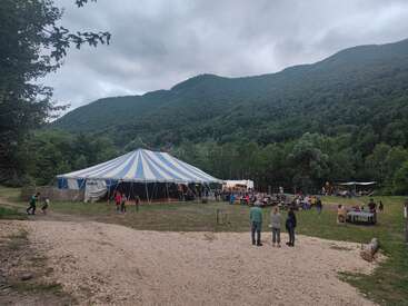 Una gran carpa a rayas azules y blancas se alza en medio de un frondoso paisaje montañoso. La gente se reúne en el exterior, sentada en las mesas, disfrutando de un evento o festival al aire libre.