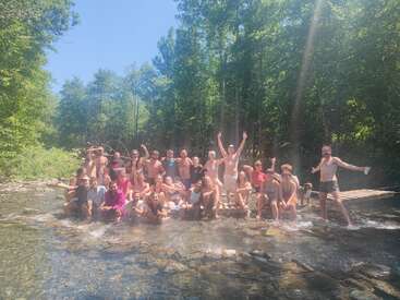 Un numeroso grupo de personas, disfrutando del sol, posa alegremente sobre una plataforma de madera en un arroyo de aguas poco profundas, rodeado de frondosos árboles y de la naturaleza.