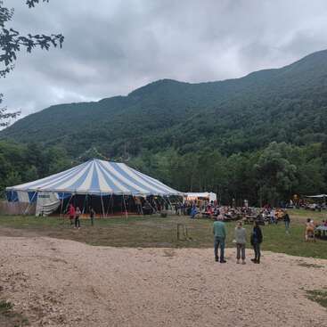 Uma grande tenda listrada de azul e branco ergue-se no meio de uma paisagem montanhosa verdejante. As pessoas se reúnem do lado de fora, sentadas às mesas, curtindo um evento ao ar livre ou um festival.