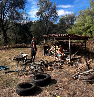 Two people are working outdoors near a rustic wood shelter, surrounded by logs, tools, wheelbarrow, and tires, under a bright sky and leafy trees.