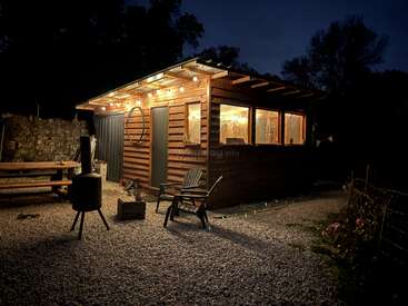 A cozy wooden cabin is warmly lit at night. Outdoor lights, chairs, a fire pit, picnic table, and trees create a peaceful, inviting atmosphere in nature.