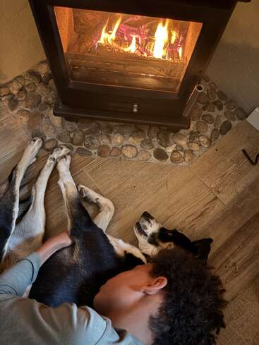 A boy lovingly cuddles with his black and white dog on a wooden floor, both relaxing in front of a warm, crackling fireplace on a cozy evening.