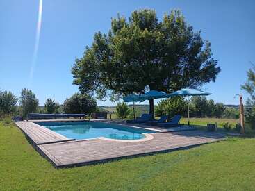 Uma piscina ao ar livre tranquila, cercada por grama verde, deck de madeira e sombreada por árvores grandes. Espreguiçadeiras azuis e guarda-sóis criam um ambiente relaxante e convidativo.