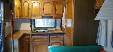 Dieses Bild zeigt das Innere einer gemütlichen Wohnmobilküche mit Holzschränken, einem Herd, einem Fenster, Arbeitsfläche, Spüle, grünem Sofa und Vorhängen.