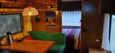 This cozy camper interior features a green cushioned dining nook, wooden table, woven lamp, colorful wall hanging, and a bed with striped bedding in the background.