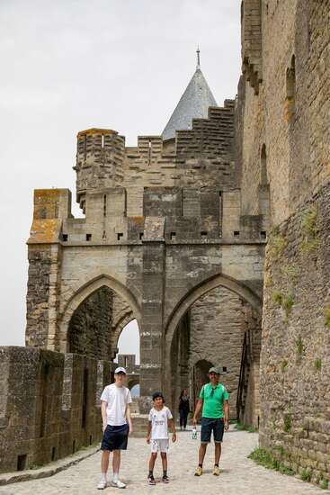 Trois personnes, deux adultes et un enfant, se trouvent sur un chemin de pierre à côté des murs d'un château médiéval et d'une porte voûtée, sous un ciel nuageux, en train d'explorer le site historique.