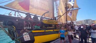 Un grand voilier historique à la coque jaune et noire est amarré dans un port très fréquenté. De nombreuses personnes sont rassemblées autour, profitant de la journée ensoleillée et des festivités.