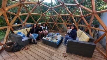 Cinq personnes sont assises à l'intérieur d'un dôme géodésique en bois sur des canapés en osier, entourés d'arbres. Elles dégustent ensemble de la nourriture et des boissons sur une petite table.