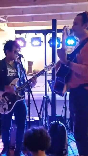 Deux musiciens se produisent en intérieur, l'un jouant de la guitare électrique et chantant, l'autre de la guitare acoustique. Les lumières colorées de la scène les éclairent, créant une atmosphère musicale vibrante.