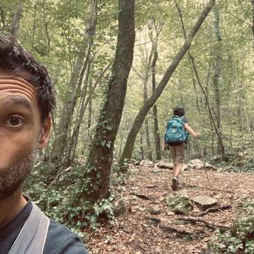 Un hombre se hace un selfie mostrando la mitad de su rostro sorprendido, mientras un niño con una mochila camina por delante a través de un frondoso y verde sendero forestal. Naturaleza tranquila.
