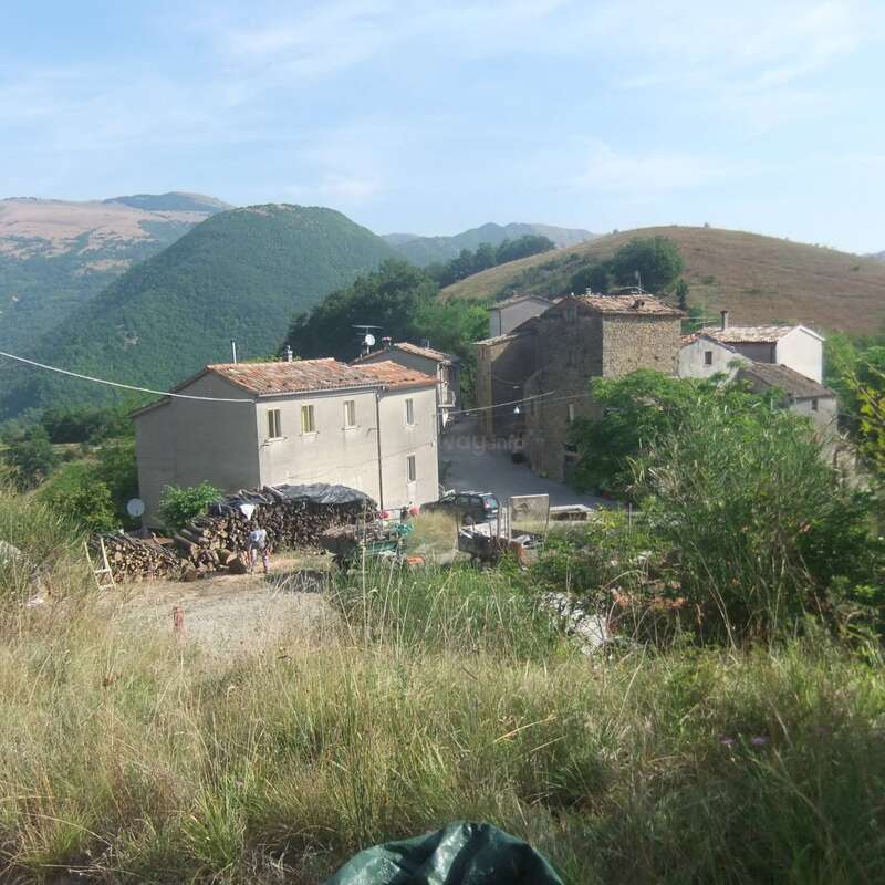 L'image représente une scène rurale sereine avec un groupe de maisons, un tas de bois de chauffage et un champ herbeux, sur fond de collines et de montagnes ondulantes.