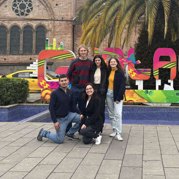 Five people pose together in front of a colorful "Cuenca" sign, with a palm tree, historical church, and yellow car in the background on a cloudy day.