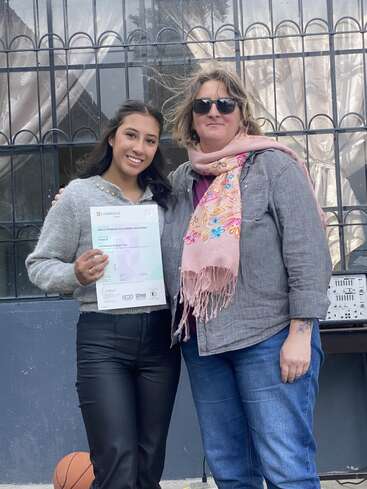 Dos mujeres sonríen juntas. Una sostiene un certificado de Cambridge. Están al aire libre, frente a una ventana con barrotes de hierro y cortinas detrás.