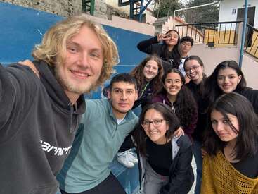 Un grupo de diez jóvenes posa para un alegre selfie en unas gradas azules, sonrientes y aparentemente felices, con ropa informal y en un ambiente relajado al aire libre.