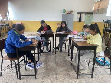 Cinco estudiantes sentados en pupitres separados, concentrados en tareas de escritura en un aula. El aula tiene paredes amarillas, un armario y sillas apiladas al fondo.