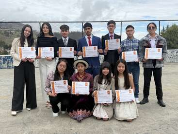 Un groupe de douze jeunes étudiants posent en plein air, tenant fièrement leurs certificats. Ils sont vêtus de manière formelle, souriants, et célèbrent leur réussite, debout ou agenouillés sur une surface en béton.