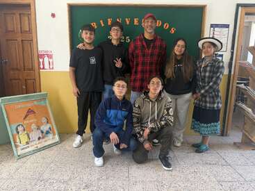 Sept personnes posent à l'intérieur d'une école, debout devant un panneau vert « Bienvenidos ». Deux sont assises par terre, les autres sont debout. Une femme porte un costume traditionnel.