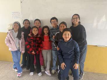 Un groupe de huit enfants souriants et un adulte se tiennent ensemble dans une salle de classe, posant pour une photo devant un tableau blanc. Leurs expressions sont joyeuses.