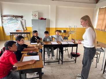 Un profesor está interactuando con un grupo de alumnos pequeños en el aula. Los alumnos están atentos, sentados en sus pupitres con sus cuadernos y un títere de juguete.