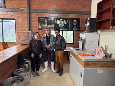 Tres personas están juntas dentro de una cervecería con paredes de ladrillo. Se ven barriles de cerveza, una barra y el equipo de elaboración de cerveza. Detrás de ellos cuelga un cartel que dice «La Cascada».