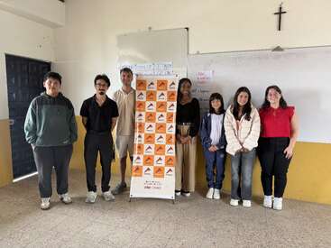 Siete personas posan en un aula junto a una pancarta, sonriendo a la cámara. Hay pizarras blancas con notas escritas a mano y una cruz en la pared.