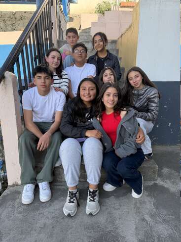 Um grupo de nove adolescentes está sentado bem juntinho numa escadaria ao ar livre, sorrindo para a câmera. Eles parecem felizes e relaxados, curtindo a companhia uns dos outros.