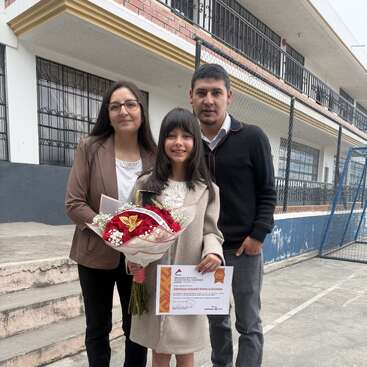 A young girl stands proudly with a certificate and bouquet, flanked by two adults, likely her parents, smiling outside what appears to be a school building.