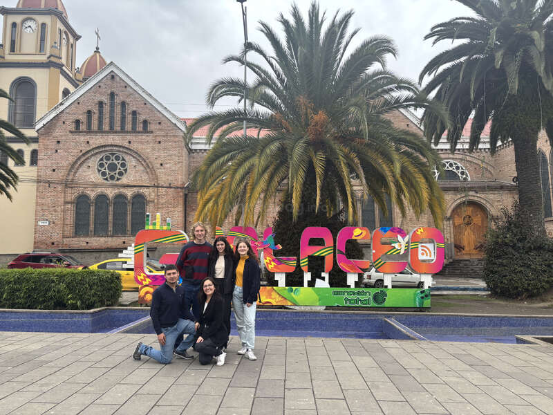 Cinco personas posan juntas delante de un colorido cartel de "Cuenca", con una palmera, una iglesia histórica y un coche amarillo de fondo en un día nublado.