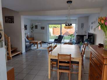 This image shows a spacious, modern living and dining area with wooden furniture, tile floors, large windows, plants, a cozy sofa, and a vase of tulips.