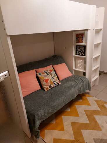 This image shows a cozy room with a white loft bed above a sofa. The sofa has colorful pillows, and shelves with books and artwork.