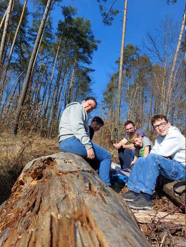 Five people sit on or near a fallen log in a sunny forest. Tall trees surround them, and everyone appears relaxed, enjoying a moment outdoors together.