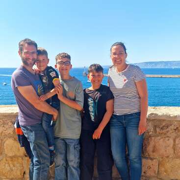 Una feliz familia de cinco miembros posa frente a un muro de piedra con el océano y las lejanas colinas a sus espaldas, disfrutando de un soleado día juntos al aire libre.