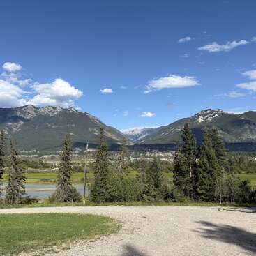 A bright, clear day showcases majestic mountains, scattered clouds, lush green trees, a winding river, and gravel path, creating a peaceful, natural landscape under a vivid blue sky.