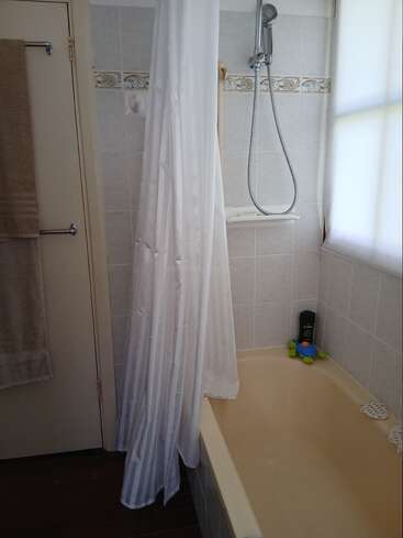 This image shows a bathroom with a cream bathtub, white shower curtain, towel racks with towels, handheld shower head, window, kid’s toy turtle, and toiletries.