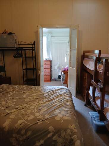 This image shows a cozy bedroom with a double bed, bunk beds, a rack, open French doors leading to a bright room with a dresser and fan.