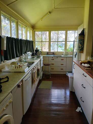 This kitchen features large windows, wooden floors, white cabinets, a dish rack, appliances, green curtains, and natural light, creating a cozy, lived-in and inviting space.