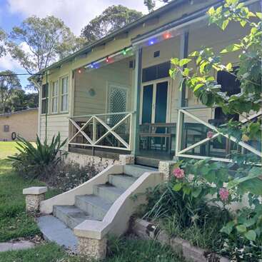 Uma casa de campo pitoresca com uma pequena varanda, luzes decorativas e um jardim simples. Degraus levam até a entrada, cercada por vegetação, flores cor-de-rosa e árvores grandes.