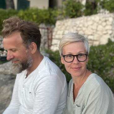 A smiling woman with short blonde hair and glasses sits next to a bearded man in white, outdoors, with greenery and a stone wall behind them.