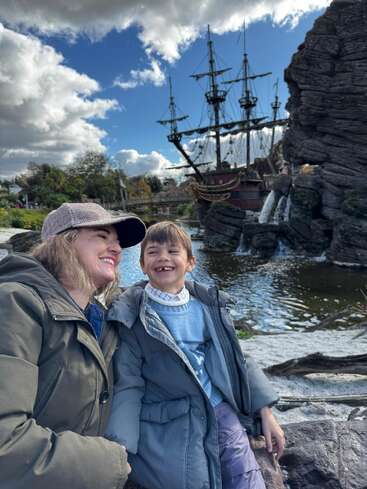 A woman and a child share a joyful moment outdoors, dressed warmly. Behind them is a large pirate ship, rocky landscape, water, and dramatic cloudy sky.