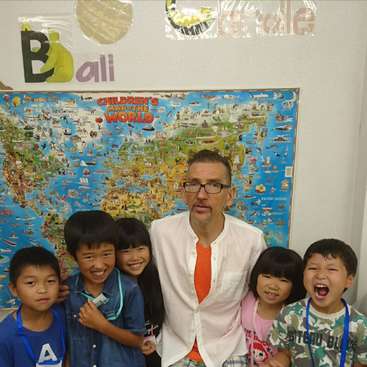 La imagen muestra a un hombre con camisa blanca posando con cuatro niños delante de un colorido mapa del mundo en una pared, con la palabra "Bali" visible encima.