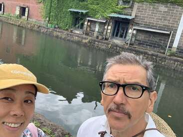 Un hombre y una mujer posan junto a un tranquilo canal, edificios históricos con hiedra al fondo, ambos aparecen relajados, con ropa informal y sombreros, disfrutando de su paseo.