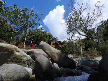 The image depicts a serene scene of a house nestled among trees and large rocks, with a small stream flowing through the foreground.