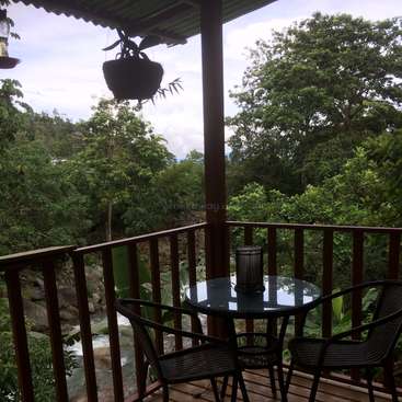 The image depicts a serene balcony with a small table and chairs, overlooking a tranquil stream and lush greenery, set against a cloudy sky.